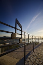 A large metallic musical note sculpture is set against a scenic background with a clear blue sky. The structure resembles a musical stave with notes and is positioned on a paved walkway with a view over a cityscape. The sun is low, casting long shadows and creating a serene atmosphere.