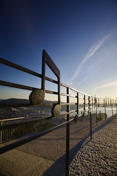 A large metallic musical note sculpture is set against a scenic background with a clear blue sky. The structure resembles a musical stave with notes and is positioned on a paved walkway with a view over a cityscape. The sun is low, casting long shadows and creating a serene atmosphere.