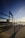 A large metallic musical note sculpture is set against a scenic background with a clear blue sky. The structure resembles a musical stave with notes and is positioned on a paved walkway with a view over a cityscape. The sun is low, casting long shadows and creating a serene atmosphere.