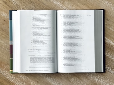 An open book is lying on a wooden surface, showcasing text on both visible pages. The book appears to have multiple colored tabs or dividers on the left side. The layout of the text suggests it is a reference or religious text with numbered verses.