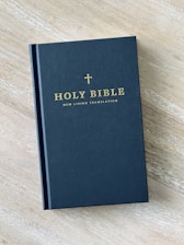 A hardcover book titled 'Holy Bible: New Living Translation' with gold lettering and a small cross above the text. The book is placed on a light wooden surface, and its cover is dark in color.