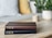 Two hardcover books are stacked neatly on a light wooden table. One book has a dark brown cover, and the other has a black cover. Both are titled 'Holy Bible NLT' and published by Tyndale. In the background, there is a blurred view of a cozy living space with a striped pillow, a yellow pillow, a potted plant with green leaves, and a small cup.
