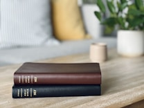 Two hardcover books are stacked neatly on a light wooden table. One book has a dark brown cover, and the other has a black cover. Both are titled 'Holy Bible NLT' and published by Tyndale. In the background, there is a blurred view of a cozy living space with a striped pillow, a yellow pillow, a potted plant with green leaves, and a small cup.