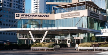 A modern hotel entrance with glass architecture and reflective surfaces. The building features a sign reading 'Wyndham Grand Istanbul Levent' and a restaurant named 'Car&ecirc;me Restaurant'. The setting includes landscaped plants and seating in the foreground, with a clear blue sky and surrounding tall buildings visible in the background.