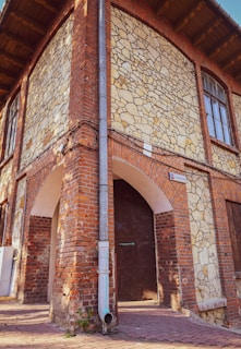 A corner of an old brick and stone building with arched doorways and a sign reading '16 Grabancza'. The facade features a mix of red bricks and irregularly shaped stones, with an exposed water pipe running down the corner. Large wooden doors are set within the arches, and a window is visible above.