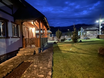 A cozy house with a well-lit wooden porch and stone steps leading to a paved walkway. The yard is spacious with neatly trimmed grass and small trees. The sky is dark with a vibrant blue hue, suggesting it's either dusk or early evening. In the background, there are a few other buildings and mountains visible.