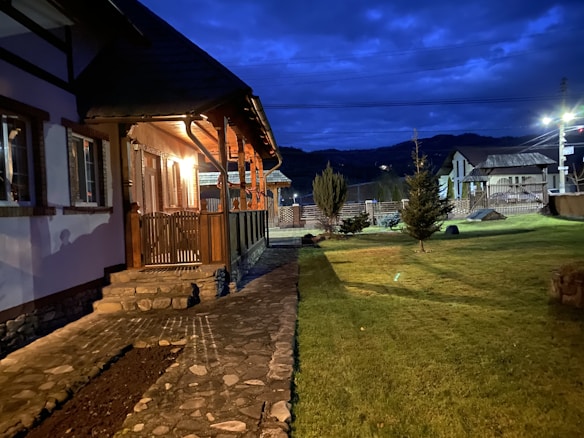 A cozy house with a well-lit wooden porch and stone steps leading to a paved walkway. The yard is spacious with neatly trimmed grass and small trees. The sky is dark with a vibrant blue hue, suggesting it's either dusk or early evening. In the background, there are a few other buildings and mountains visible.