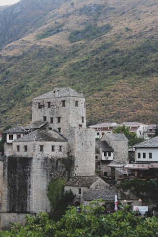 A stone fortress with multiple rectangular and cylindrical towers is set against a backdrop of a hill covered with sparse vegetation. The fortress has a medieval appearance with weathered stone walls and small, square windows. Surrounding the structure are a few residential buildings, and in the foreground, there are green shrubs and foliage.