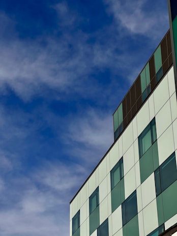 A modern building with a grid-like facade of green and white panels, set against a backdrop of a bright blue sky with scattered clouds. The building features several windows and has a sleek, contemporary design.