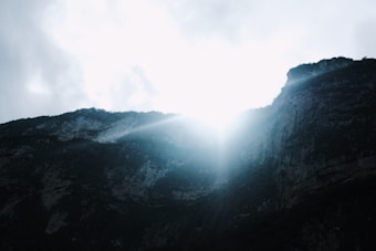 Sunlight breaks over a rugged mountain ridge, casting light across a rocky cliff. The sky is partly cloudy, giving contrast to the bright sunlight and the dark shadows on the mountain.