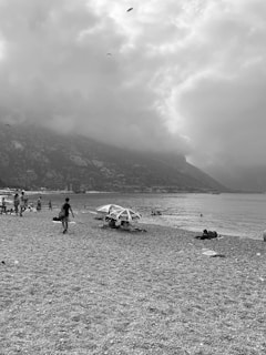 A pebble beach with a few people walking and lounging under umbrellas. The sea is calm, with some swimmers visible. Paragliders are in the distance against a backdrop of large clouds and mountainous terrain, creating a dramatic atmosphere.