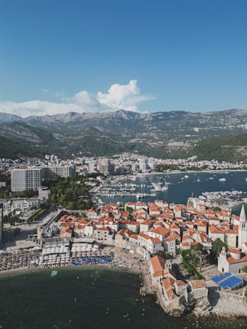 A coastal town with a dense cluster of traditional Mediterranean buildings featuring orange-tiled roofs and stone walls. The town is surrounded by a calm bay filled with numerous docked boats and yachts. In the background, a range of lush, green mountains rises under a clear blue sky. A crowded beach with sun umbrellas is visible along the coastline, indicating a popular tourist destination.