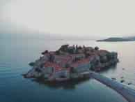 An island connected to the mainland by a narrow isthmus, featuring numerous stone buildings with red rooftops. The area is surrounded by calm blue waters, and there are a few small boats anchored nearby. Lush greenery is scattered between the buildings, providing contrast against the architectural structures and the sea.