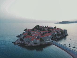 An island connected to the mainland by a narrow isthmus, featuring numerous stone buildings with red rooftops. The area is surrounded by calm blue waters, and there are a few small boats anchored nearby. Lush greenery is scattered between the buildings, providing contrast against the architectural structures and the sea.