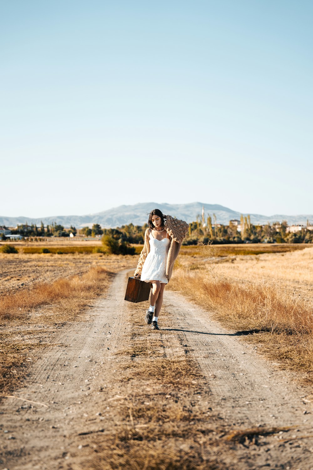 a woman walking down a dirt road carrying a suitcase