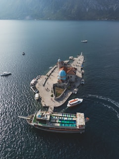 Aerial view of a small, picturesque island with a prominent blue-domed church. The island is surrounded by calm blue waters and a few boats are docked nearby. A large ferry with a colorful deck is prominently featured in the foreground. The background consists of distant misty mountains.