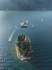 An aerial view of a small, lush island with a historic building surrounded by tall, dense trees and partially enclosed by a stone wall. Several boats, including a larger one near a nearby structure, navigate the calm blue waters. The backdrop includes distant mountains shrouded in mist.