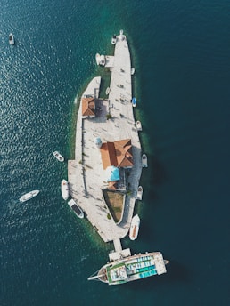 Aerial view of a small island with white stone pathways, surrounded by deep blue water. Several boats are moored around the island, and there is a structure with reddish-brown roofs, including one with a dome. People can be seen walking along the paths.