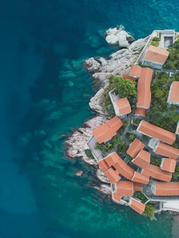 Aerial view of a cluster of buildings with red-tiled roofs set against rocky cliffs and surrounded by the turquoise blue sea. The vivid contrast between the earthy tones of the architecture and the vibrant ocean creates a visually striking scene. Lush green vegetation is interspersed among the buildings.