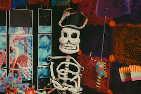A detailed setup featuring a smiling skeleton figure adorned with a black hat, situated amidst colorful, intricate papel picado hanging decorations. The backdrop showcases vivid hues, including orange and purple, with traditional patterns and symbols. Close by, a multicolored dragon or serpent poster adds to the vibrant atmosphere, while other small decorative elements, like marigold flowers, complete the scene.