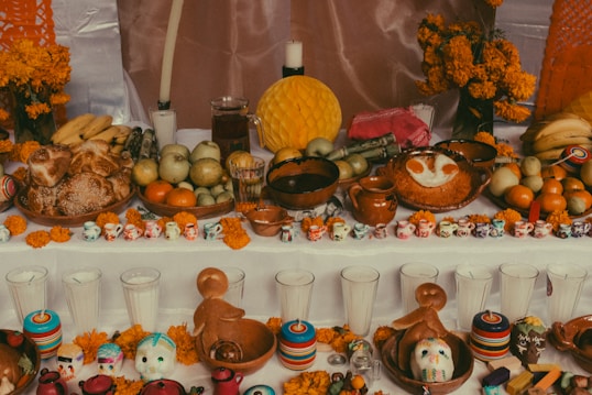 A traditional altar is decorated with vibrant marigold flowers, an assortment of fruits such as bananas and oranges, and various bread and pastries. There are small colorful skull ornaments and miniature ceramic cups arranged neatly. Clay pots and a pitcher are situated among the offerings, with candles and a decorative paper backdrop enhancing the display.