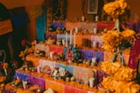 An altar decorated with vibrant papel picado, candles, marigold flowers, and various offerings such as fruit, bread, and decorated skulls. Three tiers hold these colorful arrangements, often used in traditional celebrations.