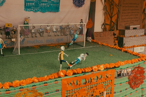 A decorated scene featuring a miniature soccer field with skeleton figures as players. These skeleton figures are engaged in a soccer match, with some in action poses, while others are seated as spectators. The area is adorned with traditional Mexican papel picado and marigold flowers, indicating a Day of the Dead theme. The backdrop displays the word 'Ofrendas' and other decorations related to the celebration.