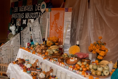 An altar is adorned with colorful decorations, including marigold flowers, papel picado, and various offerings like fruits, bread, and candles. Small skulls are placed around, and a poster remembers beloved pets.