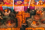 An intricately arranged altar features vibrant marigold flowers, candles, colorful papel picado, various fruits, corn, and pottery items. The central focus is a cross made of marigolds surrounded by different types of corn and seeds.