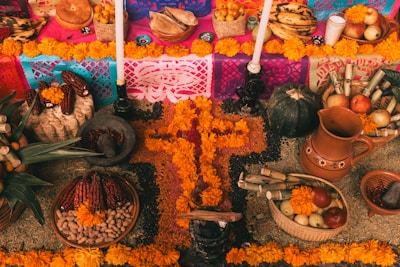An intricately arranged altar features vibrant marigold flowers, candles, colorful papel picado, various fruits, corn, and pottery items. The central focus is a cross made of marigolds surrounded by different types of corn and seeds.