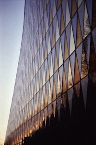 The image features the facade of a modern building with a grid of large triangular glass panels that reflect the sky and surrounding environment. The glass displays a blend of warm colors from the sunset, creating a dynamic pattern with the angular shapes.