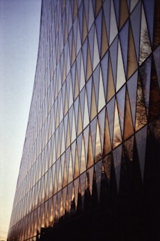 The image features the facade of a modern building with a grid of large triangular glass panels that reflect the sky and surrounding environment. The glass displays a blend of warm colors from the sunset, creating a dynamic pattern with the angular shapes.