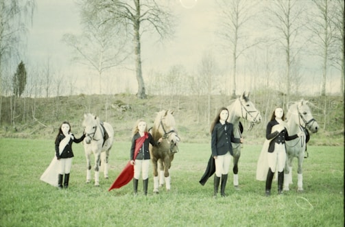 Four people in equestrian attire stand alongside horses on a grassy field. They are dressed in dark blazers, white pants, and are holding the reins of their horses. The scene is set against a backdrop of bare trees and overcast skies, creating a serene and calm atmosphere.