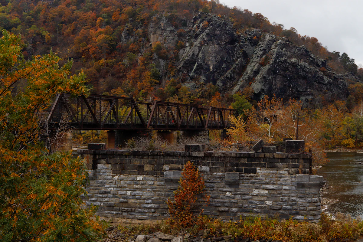An iron railroad bridge and a stone pier at Harpers Ferry, below a rocky cliff in fall color
