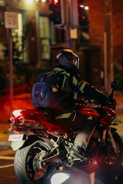 A motorcyclist wearing a helmet and a black and green jacket rides a red sports motorcycle. A blue backpack is strapped to the rider's back. The background is urban, with blurred elements suggesting evening or night lighting. Traffic lights and street signs are visible.