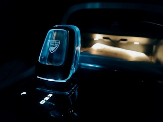A close-up view of a modern car gear shifter with illuminated letters P, R, N, and D indicating gear positions. The shifter has a sleek design and features a logo, 'Orrefors Sweden.' The background is dimly lit, showcasing part of the car's dashboard with ambient lighting.