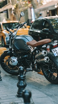 A stylish motorcycle with a brown seat and large exhaust pipes is parked on a cobblestone street. It is surrounded by black safety bollards and urban vehicles including a yellow car, suggesting a city setting.