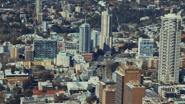 A dense urban landscape with numerous high-rise buildings of varying architectural styles. Among the structures, prominent signage such as 'Coca-Cola' and 'HARDERS' is noticeable. The area appears to be a mix of residential and commercial buildings surrounded by patches of greenery.