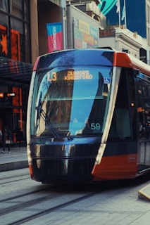 A modern tram is traveling through a city street with buildings and pedestrians visible in the background. The tram is marked 'L3 JUNIORS KINGSFORD' and is designed with sleek, reflective surfaces. Urban architecture and commercial signs are present around the tram.