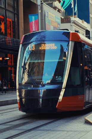 A modern tram is traveling through a city street with buildings and pedestrians visible in the background. The tram is marked 'L3 JUNIORS KINGSFORD' and is designed with sleek, reflective surfaces. Urban architecture and commercial signs are present around the tram.