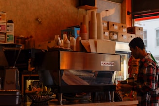 The image depicts the interior of a coffee shop. A person in a plaid shirt stands next to a large espresso machine, placing a lid on a cup. Stacks of paper cups, napkins, and various food items are arranged on top of the espresso machine. Shelves filled with boxes and coffee-related supplies line the background. A basket of bananas is visible on the counter next to the espresso machine.