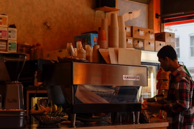 The image depicts the interior of a coffee shop. A person in a plaid shirt stands next to a large espresso machine, placing a lid on a cup. Stacks of paper cups, napkins, and various food items are arranged on top of the espresso machine. Shelves filled with boxes and coffee-related supplies line the background. A basket of bananas is visible on the counter next to the espresso machine.