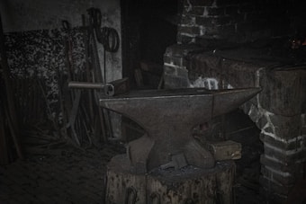 An old anvil is positioned on a sturdy wooden stump in a dimly lit workshop. Surrounding the anvil are various blacksmithing tools, including hammers and tongs, leaning against a brick wall. The background appears to be a traditional forge area with a rustic ambiance, featuring a brick hearth.