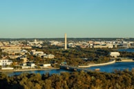 Aerial view of a city with notable landmarks including an obelisk-like monument and several prominent buildings surrounded by lush greenery and a river. The scene captures a built-up area interspersed with trees and water, under a clear blue sky.