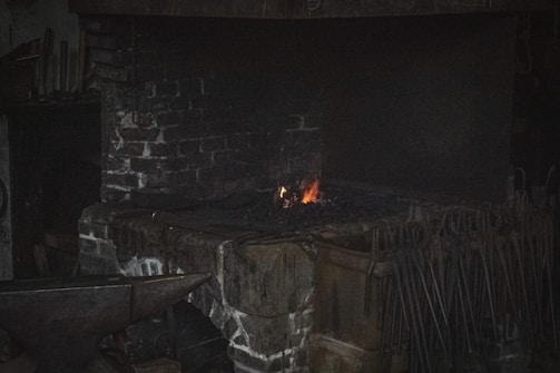 A dimly lit blacksmith's forge with a small fire glowing on a brick hearth. An anvil and various tools hang nearby, creating a rustic and industrial atmosphere.