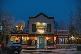 A nighttime scene of a festive building decorated with Halloween-themed decorations, including large illuminated pumpkin lanterns and a giant skull on the roof. The building, identified as 'Fatty's Pizzeria & Pasta', is covered in snow, contributing to a wintry and eerie atmosphere. Trees with snow-covered branches surround the building.