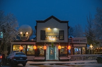 A nighttime scene of a festive building decorated with Halloween-themed decorations, including large illuminated pumpkin lanterns and a giant skull on the roof. The building, identified as 'Fatty's Pizzeria & Pasta', is covered in snow, contributing to a wintry and eerie atmosphere. Trees with snow-covered branches surround the building.