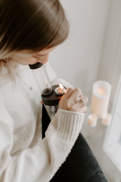A person is using a glass bong, holding a lighter to ignite the contents. The setting appears cozy, with a soft sweater and candles nearby creating a warm ambiance.