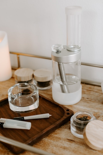 A wooden tray on a table holds a rolled cigarette and a lighter. Next to it is a glass ashtray and a few small jars with wooden lids containing different substances. A sleek, modern smoking device with clear and metal parts is also present.