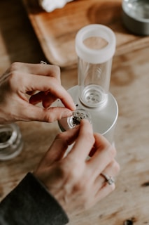 A person's hands are placing a small amount of dried herbal material into a glass chamber on a metallic device. In the background, there is a wooden tray and some other objects slightly out of focus.